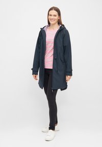 Giacca impermeabile blu navy con cappuccio, dotata di cordini regolabili e tasche laterali. Indossata sopra una camicia a righe bianche e rosa e jeans neri.