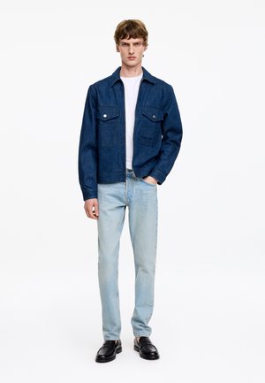 Junger Mann trägt eine dunkelblaue Jeansjacke, weißes T-Shirt, hellblaue Jeans und schwarze Leder-Slipper, steht vor weißem Hintergrund.