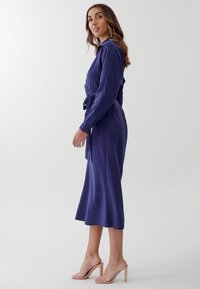 Abito midi blu royal con maniche lunghe, legato in vita, caratterizzato da una trama liscia. Indossato con sandali con tacco nudo.