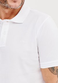 Biała koszulka polo z kołnierzykiem, z zapięciem na guziki i subtelnym haftowanym logo na piersi. Gładki, teksturowany materiał.