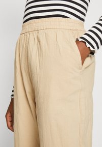 Pantalons larges beiges avec une taille élastique et des poches latérales, fabriqués dans un tissu doux et texturé. Un haut rayé noir et blanc est partiellement visible.