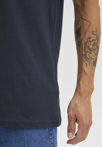 Tamsiai mėlynas medvilninis marškinėliai trumpomis rankovėmis, su šoninėmis skeltuvėmis ir maža juoda etikete prie žemyn, derinami su džinsais ir tatuiruota ranka.