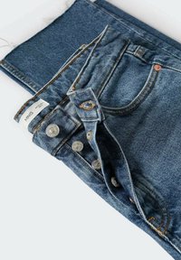 Jean en denim bleu plié avec braguette à boutons apparents, poches avant et surpiqûres orange sur fond blanc.