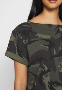 T-shirt camo vert olive avec des manches retroussées, fabriqué en coton doux. Présente une coupe décontractée et un large col, avec des motifs verts foncés et clairs.