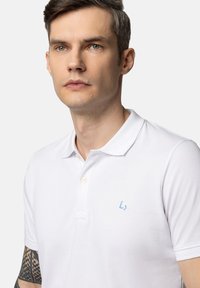 Biała koszulka polo wykonana z bawełny, z dwoma guzikami przy zapięciu, krótkimi rękawami i subtelnym niebieskim logo na piersi. Gładka tekstura.