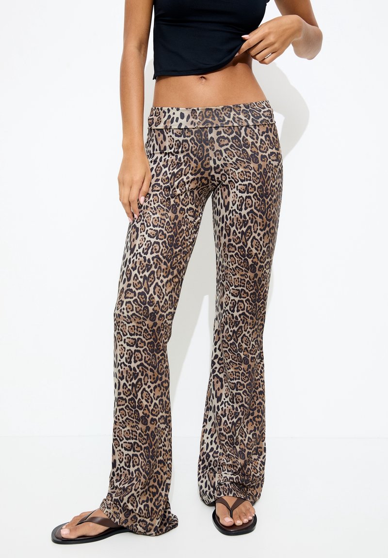 Femme portant un pantalon large taille haute à imprimé léopard, un crop top noir et des tongs noires sur un fond blanc.