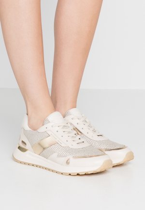 Beine in beige und goldenen Michael Kors Sneakers mit Mesh, Lederdetails und weißen Gummisohlen auf einem hellgrauen Boden.
