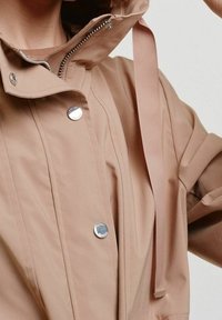 Beige voděodolná bunda s vysokým límcem, stříbrnými patentkami a předním zipem. Má strukturovanou látku a nastavitelné šňůrky na kapuci.