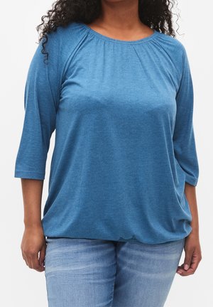 Kobieta ubrana w luźną, niebieską bluzkę z rękawami do łokcia oraz jasnoniebieskie jeansy, widoczna od ramion do bioder na jednolitym tle.