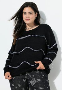 Schwarzer Pullover mit weißen Wellenstich-Details, langärmlig und mit lockerer Passform. Kombiniert mit schwarzen Jeans im Sternmuster.