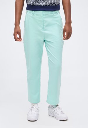 Személy világos türkiz egyenes szárú nadrágot, fehér Adidas tornacipőt és kék mintás felsőt visel, fehér háttér előtt áll.