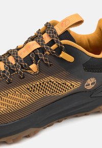 Black and orange hiking shoe with textured upper, patterned detailing, and rubber sole. Features black laces and a tree logo on the side.