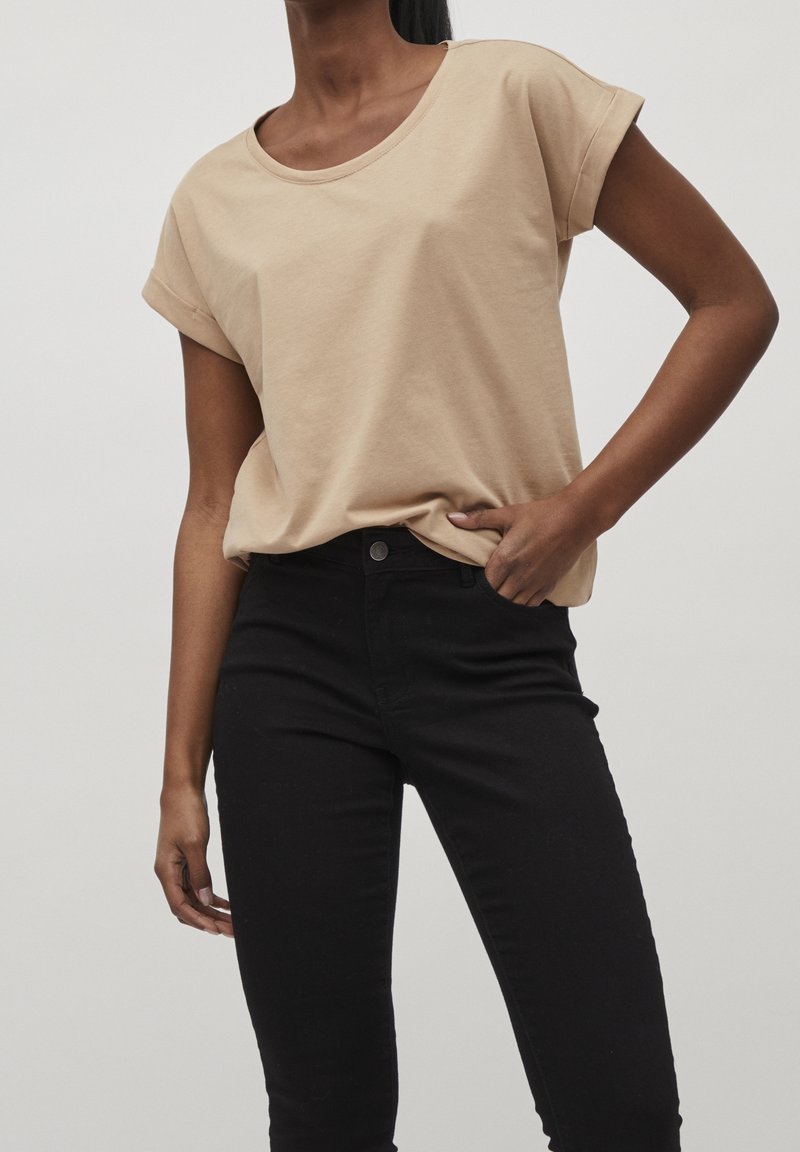 T-shirt en coton beige avec un col rond et des manches courtes roulées, associé à un jean skinny noir taille haute. Design simple et décontracté.