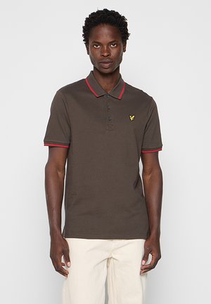 Mand iført brun poloshirt med rød kant og gul logo, kombineret med off-white bukser, stående mod en ensfarvet lys baggrund.