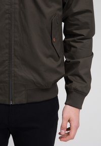 Detailní záběr na trup osoby, která má na sobě tmavě zelenou bundu se zipem, kapsou na knoflík a žebrovanými manžetami, stojící na světlém pozadí.