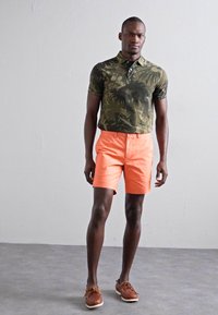 Polo cu mânecă scurtă în verde măsliniu cu model de frunze, asortat cu pantaloni scurți portocalii strălucitori și pantofi de bărbat maro. Material neted, casual.
