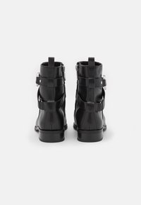 Une paire de bottines en cuir noir avec des boucles sur les côtés et des tirettes à l'arrière, vues de l'arrière sur un fond clair.