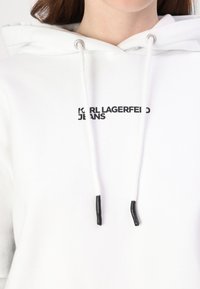 Hanorac alb cu cordon, având capse negre. Include inscripția "KARL LAGERFELD JEANS" imprimată în text negru pe față. Material moale.