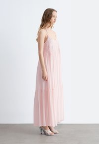 Jeune femme debout de profil, portant une longue robe sans manches rose pâle avec broderies et des sandales argentées à talons hauts.