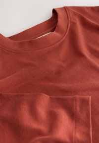 Bawełniana koszulka w kolorze rdzy, z okrągłym dekoltem, krótkimi rękawami i małą kieszonką na piersi. Gładka faktura z widocznymi detalami szwów.