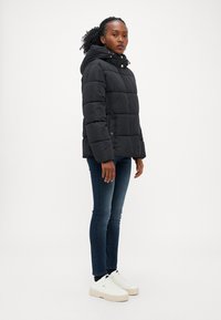 Svart boblejakke med hette, quiltet design, gullfargede detaljer, kombinert med mørkeblå jeans og hvite joggesko.