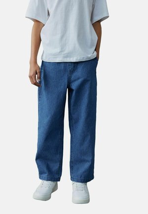 Blaue gestreifte Hose aus Denim, lockere Passform, gerade geschnittenes Bein, kombiniert mit weißen Sneakers. Einfaches weißes T-Shirt darüber getragen.