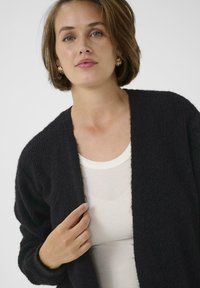 O femeie cu părul scurt, brunet, poartă cercei aurii în cerc, un top alb cu dungi și o cardigan texturată neagră, ținând marginea acesteia.