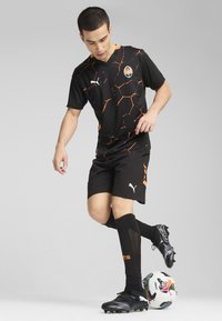 Camiseta de fútbol negra con patrón hexagonal naranja, pantalones cortos negros, botas de fútbol negras y calcetines negros con diseño de malla. Pelota de fútbol a sus pies.