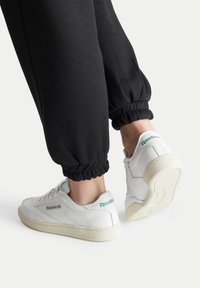 Balti odiniai sportbačiai su žaliais akcentais, guminiais padais ir perforuotais raštais. Dėvimi su juodais treniruokliais su elastinėmis apykaklėmis.