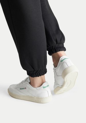 Valged nahast tenniseksid roheliste aktsentidega, kummist tallad ja perforatsioonidega detailid. Kanda mustade dressipükstega, millel on elastsed mansetid.