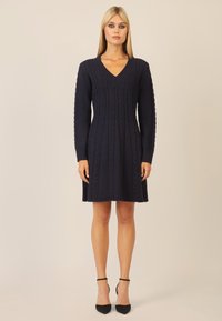 Navy gestricktes Kleid mit V-Ausschnitt und langen Ärmeln, das ein Zopfmuster, einen ausgestellt Rock und eine glatte Textur aufweist, kombiniert mit schwarzen Absätzen.
