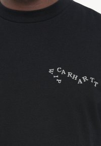 Svart bomulls-T-skjorte med rund hals, med buet hvit tekstlogo på venstre side. Myk tekstur, klassisk design.