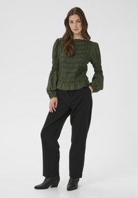 Dunkelgrüne Smocked-Bluse mit langen, puffigen Ärmeln und rüschigem Saum, kombiniert mit schwarzen, weit geschnittenen Hosen und schwarzen, hohen Stiefeln.