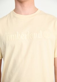 Beige katoenen t-shirt met een ronde hals. Heeft een reliëf 'Timberland' logo en een boomicoon op de voorkant. Gladde textuur, korte mouwen.