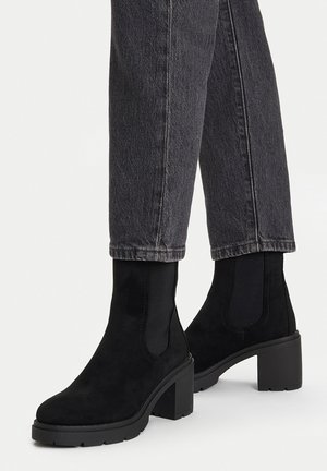 Stivaletti alla caviglia in camoscio nero con tacchi chunky indossati con jeans denim grigio scuro, mostrati su uno sfondo chiaro e uniforme.