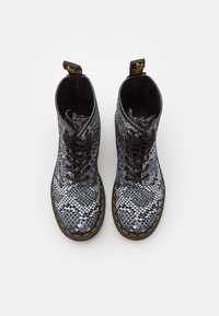 Botas de tobillo con estampado de serpiente en blanco y negro, con puntera redonda, cordones negros y costuras amarillas. La vista superior muestra las aberturas de las botas y el logotipo.