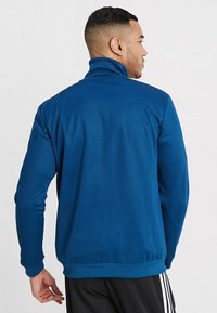 Modrá zipová bunda s vysokým límcem, vyrobená z hladké tkaniny. Má dlouhé rukávy a přiléhavý pas. Žádné viditelné vzory nebo akcenty.