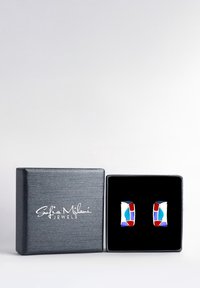 Srebrne kolczyki w geometryczne wzory w kolorze czerwonym, niebieskim i białym, wyeksponowane w teksturowanym czarnym pudełku z logo.