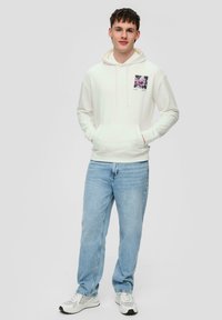 Sudadera blanca con capucha y bolsillo frontal, con un estampado gráfico cuadrado en el lado izquierdo, combinada con vaqueros de color azul claro y zapatillas.
