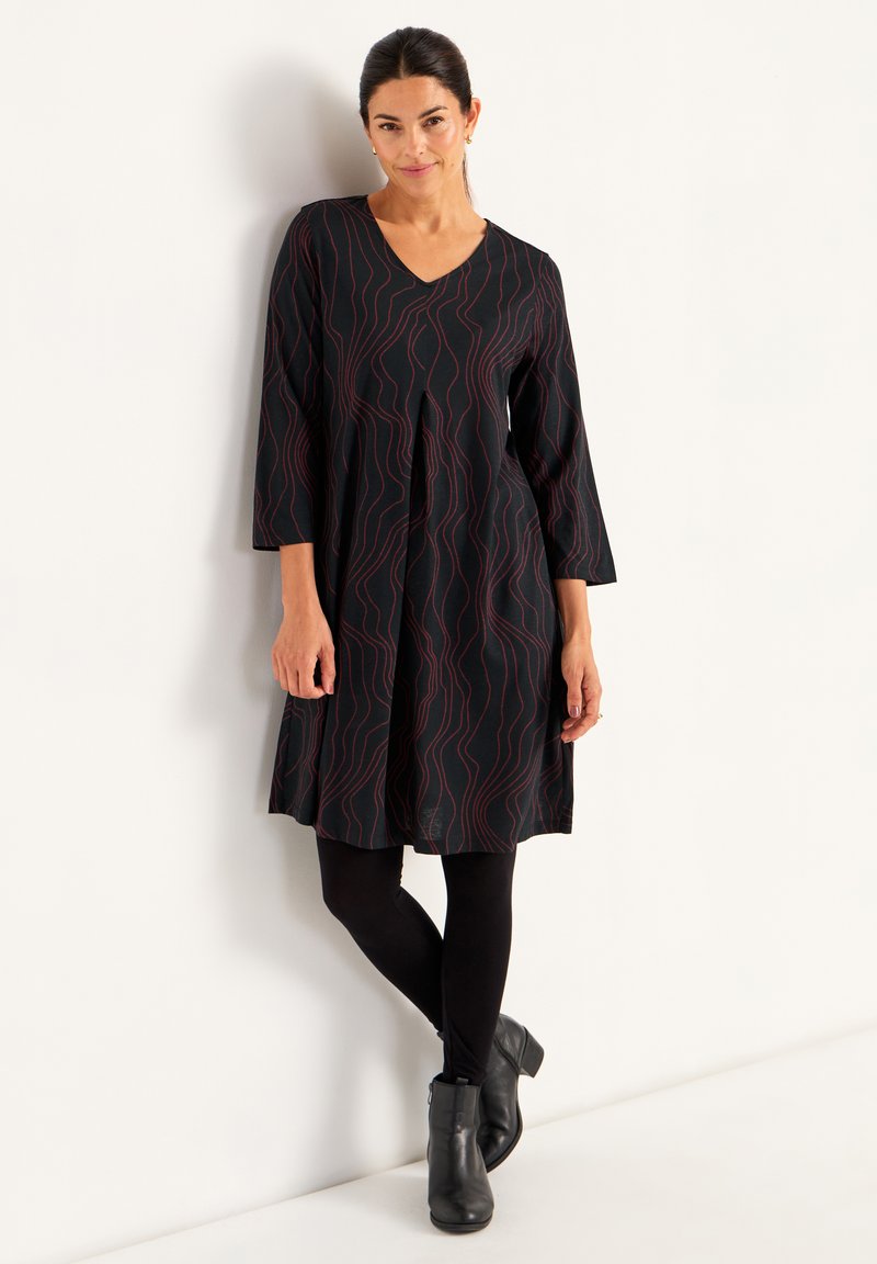 Svart knälång klänning med röd vågmönster, V-formad halsringning, 3/4 ärmar och utsvängd design, kombinerad med svarta leggings och ankelstövlar.