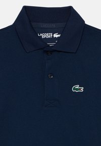 Ναυτικό μπλε polo μπλούζα της Lacoste Sport με ραμμένο γιακά, δύο μαύρα κουμπιά και ένα μικρό πράσινο λογότυπο κροκόδειλου στο στήθος.