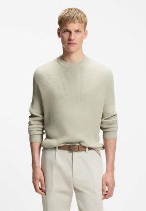 Giovane uomo con capelli biondi corti che indossa un maglione a coste beige, pantaloni chiari e una cintura marrone su sfondo bianco.