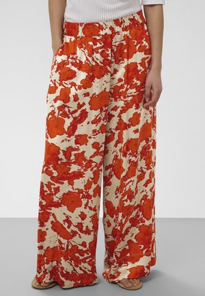 Pantalon à jambes larges avec imprimé floral abstrait rouge et crème, porté avec un haut blanc et des sandales, mains détendues le long du corps.