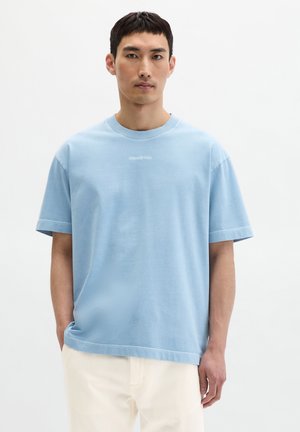 Mann trägt ein hellblaues, übergroßes T-Shirt mit kurzen Ärmeln und weiße Hose, steht vor einem schlichten weißen Hintergrund.