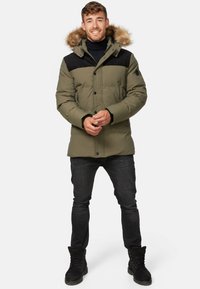 Olivenfarvet pufferjakke med sorte detaljer, pelsforet hætte, trykknapper, sidelommer, båret med sorte jeans og støvler.