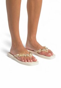 Goudkleurige flip-flops met decoratieve versieringen, voorzien van een gestructureerde crèmekleurige zool. Open teen ontwerp dat verzorgde nagels laat zien.