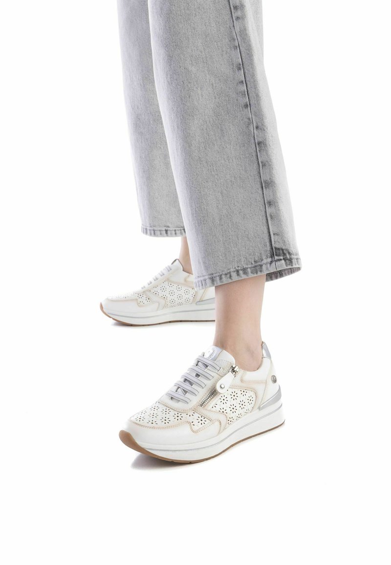 Deportivas blancas con parte superior de cuero perforado, acentos grises, cordones y un cierre lateral. Suela de goma con una banda de rodadura marrón clara, diseño redondeado.