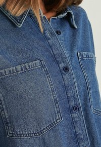 Primo piano di una camicia in denim blu con due tasche sul petto e bottoni blu scuri, indossata da una persona con capelli castano chiaro.