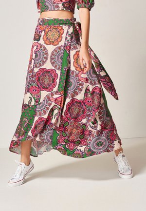 Vrouw draagt een kleurrijke wikkelrok met bloemen- en geometrisch patroon en witte sneakers, staand met gespreide benen op een effen achtergrond.
