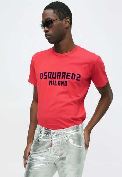 Punane puuvillane T-särk, millel on tumesinised trükitud "DSQUARED2 MILANO" tekst. Mudel kannab läikivaid hõbedaseid metallilisi teksaseid, mis rõhutavad säravat disaini.